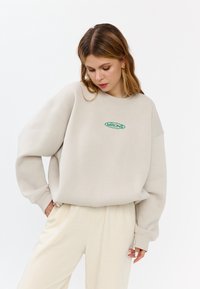 Beige katoenen oversized sweatshirt met groene geborduurde "MIRONS" logo, ribgebreide manchetten en ontspannen pasvorm, gecombineerd met crèmekleurige broeken.