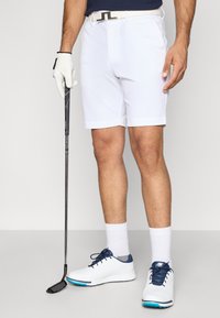 Λευκά σορτς γκολφ με υφασμάτινο σχέδιο, συνδυασμένα με μπλε μπλούζα, λευκά γάντια και λευκά παπούτσια με μπλε λεπτομέρειες. Κρατάει ένα μπαστούνι γκολφ στο χέρι.