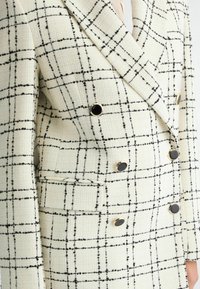 Veste croisée en tissu crème avec un motif à carreaux noirs, dotée de boutons noirs et d'un tissage texturé subtil. Deux poches avant incluses.