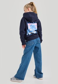 Laivastonsininen huppari, jossa on graafinen kuvitus selässä, yhdistettynä löysiin sinisiin farkkuihin ja harmaisiin lenkkareihin, joissa on valkoiset raidat. Pehmeä, rento tyyli.