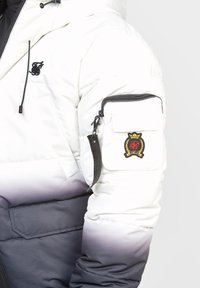 Kurtka puchowa z białą górą i ciemnoszarym dołem, z zamykaną na zamek kieszenią na rękawie i haftowanym detalem z logo. Gładka tekstura.