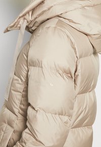 Chaqueta acolchada beige con una textura lisa y brillante. Cuenta con un cuello alto, bolsillos laterales y una marca en la manga. Capucha ajustable con cordones.