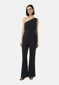 Zwarte, één-schouder jumpsuit met een ceinture taille en brede pijpen. Glad textuur met een minimalistische stijl. Model draagt hakken.
