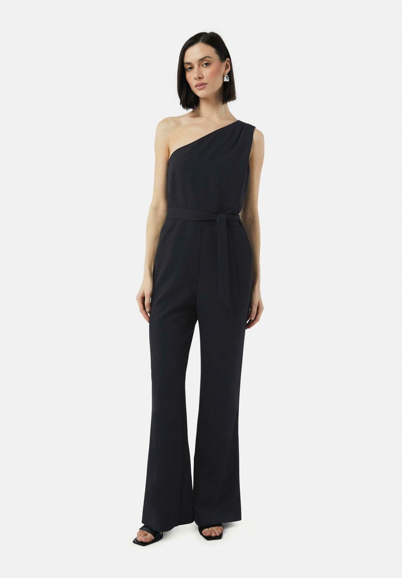 Zwarte, één-schouder jumpsuit met een ceinture taille en brede pijpen. Glad textuur met een minimalistische stijl. Model draagt hakken.