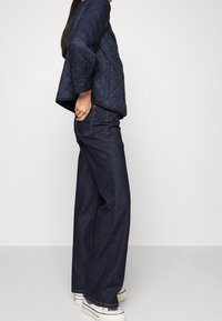Giacca trapuntata blu navy con vestibilità rilassata, abbinata a jeans a gamba larga in denim scuro a vita alta e sneakers bianche.