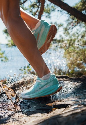 Corredor con zapatos en tonos verde azulado y blanco pisando suelo rocoso mojado cerca del agua y árboles en un entorno exterior brillante.