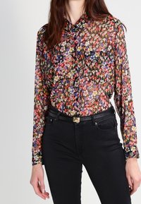 Transparent blus med ett livfullt blommönster i rött, gult, lila och blått, med en krage och en enkel bröstficka. I sällskap med svarta jeans.