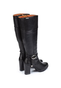 Botas negras de cuero hasta la rodilla con un eje ancho, tacón grueso, cremallera lateral y un adorno en forma de anillo cerca del tobillo. El interior presenta una tela naranja.