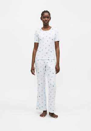 Žena stoji bosonoga, nosi bijelu majicu kratkih rukava i hlače s malim plavim cvjetnim uzorkom na bijeloj jednobojnoj pozadini.