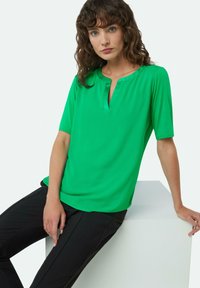 Green short-sleeve top with a keyhole neckline, smooth texture, flowing design, and paired with black slim-fit trousers.