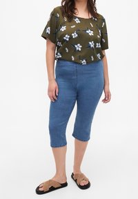 Top floral verde oliva con mangas cortas, combinado con jeans capri azules y sandalias cruzadas color tostado. La textura parece suave y casual.