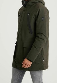 Ciemnozielona, ocieplana kurtka parka z kapturem, kieszeniami na zamek i zapięciem na napy. Teksturowany materiał z logo na rękawie.