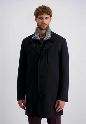 Mann trägt einen schwarzen Mantel mit grauem Kunstpelzkragen, steht vor einem schlichten hellgrauen Hintergrund und blickt leicht zur Seite.