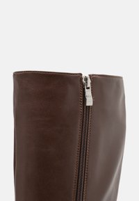 Gros plan sur une tige de botte en cuir marron foncé avec une fermeture éclair argentée visible sur le côté et des coutures soignées le long des bords.