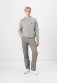 Ljust grått stickat tröja med rund hals, ihop med beige kordfluwelpants. Modellen bär vita sneakers, händerna i fickorna, neutral bakgrund.