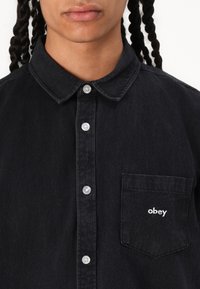 Black denim shirt with a button-up design, pointed collar, front pocket, and white embroidered "obey" logo. Textured fabric and silver buttons.