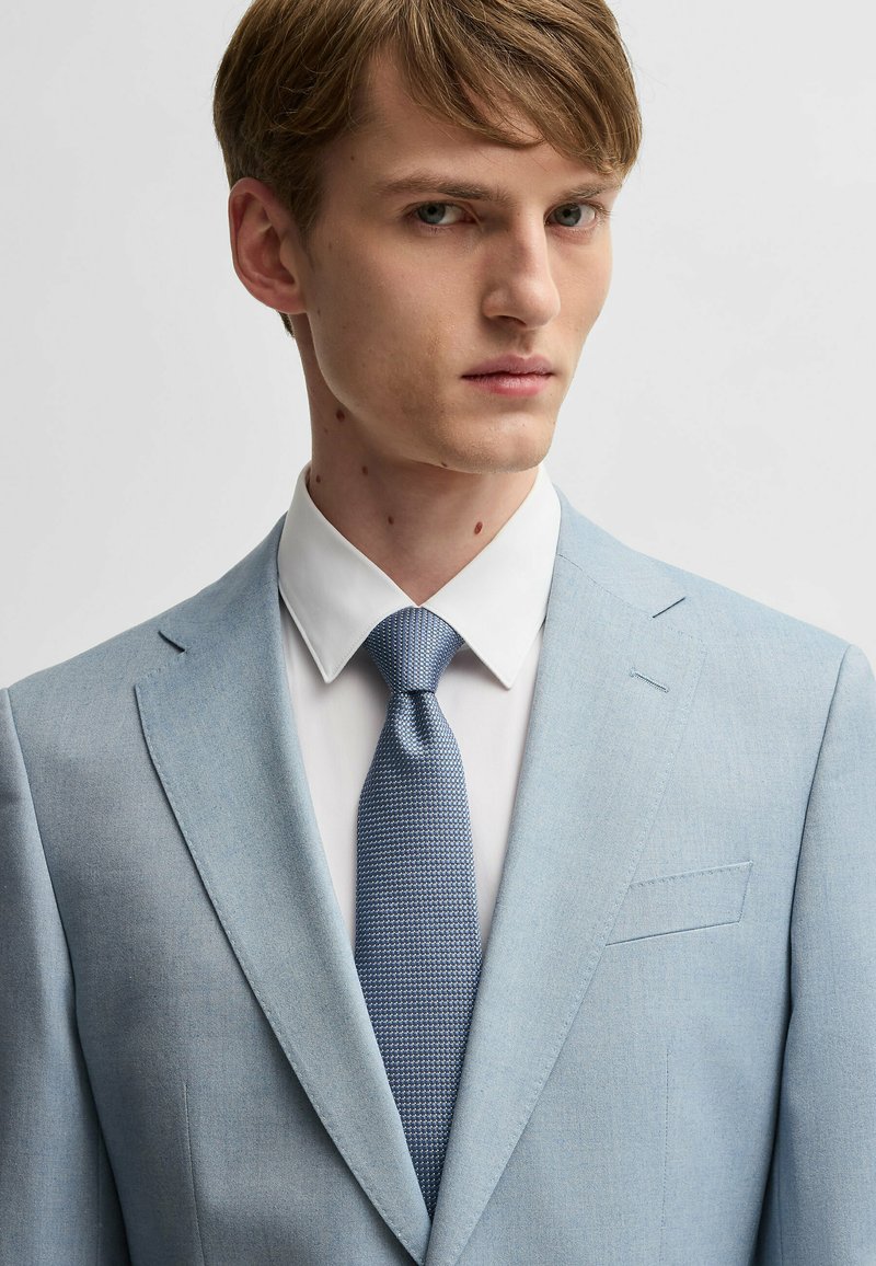 Light blue suit jacket with notched lapels and a single chest pocket, paired with a white shirt and textured blue tie. Smooth fabric finish.