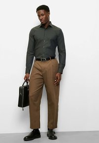 Dunkelgrünes Hemd mit Knöpfen, braune Hose, schwarzer Gürtel und Schuhe. Mann hält einen schwarzen Aktenkoffer und steht vor einem einfarbigen Hintergrund.