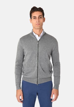 Cardigan grigio con zip realizzato in materiale morbido, dotato di un colletto alto, due tasche frontali e polsini e orlo a costine. Indossato sopra una camicia bianca.