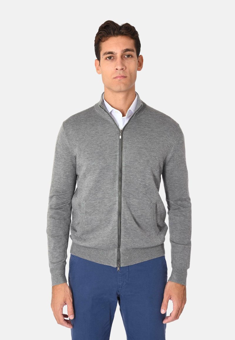 Cardigan grigio con zip realizzato in materiale morbido, dotato di un colletto alto, due tasche frontali e polsini e orlo a costine. Indossato sopra una camicia bianca.