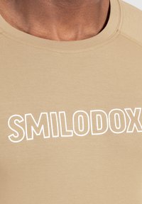 Kortärmad tröja i tan med vit text "SMILODOX" över bröstet, tillverkad av ett mjukt och lätt tyg med rund halsringning.