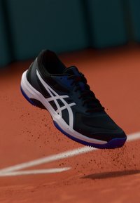 Černá sportovní bota s bílou a modrou podrážkou, s texturovaným svrškem a charakteristickým bočním logem, vznášející se nad hliněným povrchem.