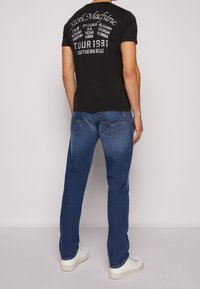 Czarna koszulka z krótkim rękawem z białym tekstem graficznym na plecach, zestawiona z niebieskimi dżinsami z lekkim wyblaknięciem i przeszyciami na kieszeniach.