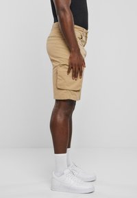 Shorts cargo beiges avec poche latérale, en coton, coupe droite. Portés avec des baskets blanches et des chaussettes à mi-mollet. Le fond est gris uni.