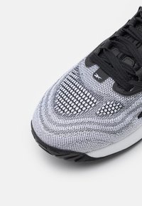 Svart och vit atletisk sko med en strukturerad stickad ovandel, snörningsstängning och en svart gummisula. Märkbar nätdetaljering nära tån.