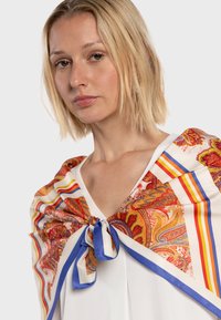 Foulard en soie arborant un motif paisley en rouge et orange, bordé de rayures bleues et jaunes, noué au cou avec un ruban bleu.