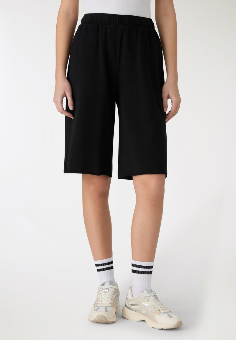 Czarne, sięgające kolan spodenki wykonane z miękkiego materiału z elastycznym paskiem w talii. Noszone z jasnymi trampkami i paskowanymi skarpetkami.