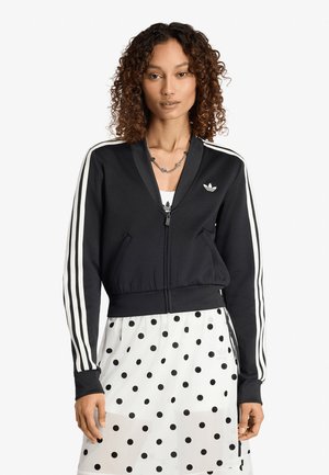 Donna che indossa una giacca Adidas nera con cerniera e strisce bianche sulle maniche e una gonna bianca con pois neri su sfondo bianco.