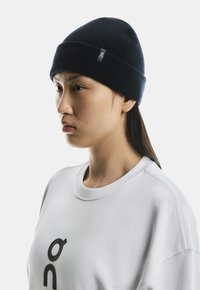 Young woman wearing a navy knit beanie and white sweatshirt with black logo, looking to the left against a plain background.