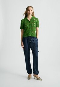 Haut vert en crochet à manches courtes avec fermeture à bouton, associé à un jogging bleu marine à taille élastique et des chaussures à talons beiges à motifs.