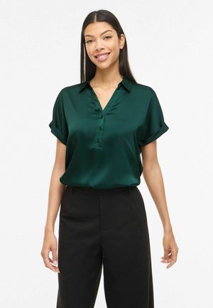 Femme souriante portant un chemisier à manches courtes boutonné vert foncé rentré dans un pantalon noir taille haute, debout devant un fond blanc uni.