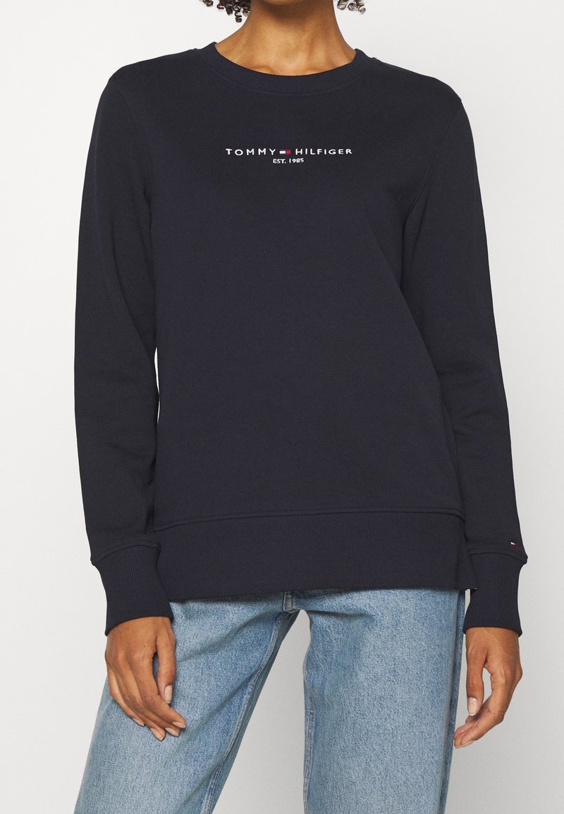 Personne portant un sweat-shirt à manches longues noir Tommy Hilfiger avec texte du logo et jean bleu clair, debout devant un fond uni.