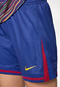 Shorts de sport bleus avec des rayures rouges sur les côtés et un logo Nike swoosh jaune, portés avec une chemise à rayures multicolores.