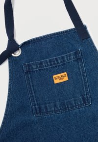 Sötétkék vászon kötény, elülső zsebbel, narancssárga címkével. Tengerészkék kötőkkel és fém szemmel az állítható illeszkedés érdekében.