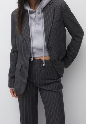 Femme portant un costume gris foncé à fines rayures avec un blazer court et un pantalon, associé à un sweat à capuche court gris clair laissant le ventre découvert.