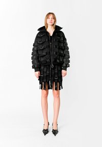 Chaqueta bomber negra con detalles de cintas en capas, combinada con una falda a juego que presenta tiras verticales, y tacones negros de punta.