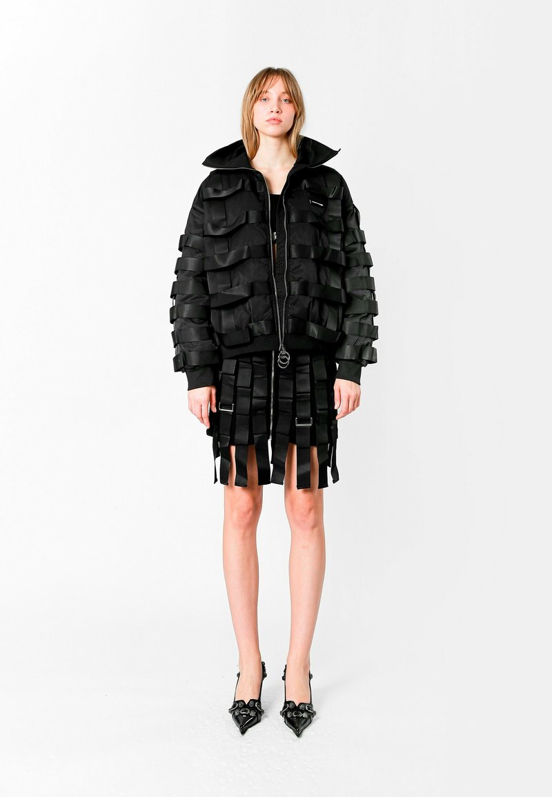 Chaqueta bomber negra con detalles de cintas en capas, combinada con una falda a juego que presenta tiras verticales, y tacones negros de punta.