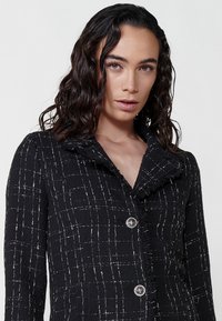 Sacou cu carouri alb-negru, dintr-un material texturat, având un guler plisat, două butoane argintii și o croială ajustată.