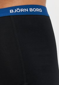 Boxer shorts noirs avec une ceinture élastique bleue portant le texte "BJÖRN BORG" en blanc. Tissu lisse et coutures plates pour un confort optimal.