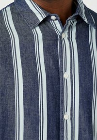 Närbild av en mörk denimskjorta med ljusblå vertikala ränder och vita knappar på en persons övre bröst och hals.