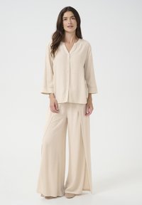 Beige skjorte med V-udskæring og trekvartærmer, parret med bredbenede beige bukser med sideslidser. Glat, let tekstur.