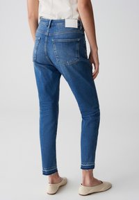 Jeans skinny en denim bleu moyen, avec une taille haute, cinq poches et un ourlet effiloché. La vue arrière montre un matériel minimal et une texture lisse.