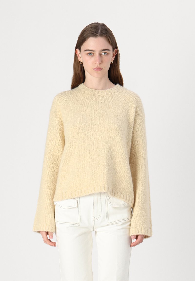 Beige gebreide trui met een ronde hals, losse pasvorm, geribbelde mouwen en een iets verkorte zoom. Draagt met witte rechte jeans.