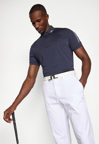 Námořnická polokošile s krátkým rukávem a límečkem, s logem na rameni, spárovaná s bílými kalhotami a bílým opaskem.