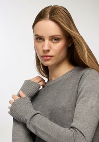 Szary sweter z dzianiny z prążkowanymi mankietami, posiadający okrągły dekolt i teksturowany wzór. Zbliżenie pokazujące ręce przy dekolcie.