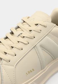 Sneaker beige con finitura in pelle liscia, dotata di lacci piatti, colletto imbottito e dettaglio del logo dorato sul lato.
